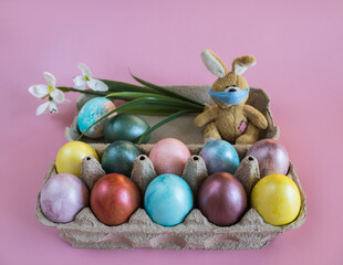 Happy Easter holiday! Fluffy Easter bunny toy in disposable  mask sits in the box with multi-colored painted eggs on pink background. Coloring at home. Easter holiday during pandemic of coronavirus