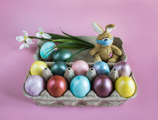 Happy Easter holiday! Fluffy Easter bunny toy in disposable  mask sits in the box with multi-colored painted eggs on pink background. Coloring at home. Easter holiday during pandemic of coronavirus