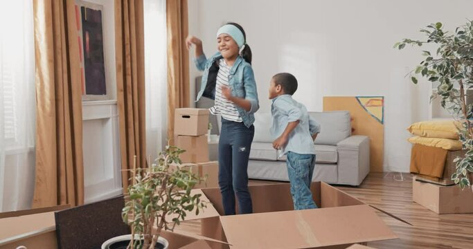 Siblings play while moving in cardboard boxes, new apartment, renovation, renting house, they hide between packed things, jump up, dancing, joy, childhood, girl takes care of younger boy