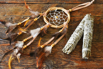 A close up image of a hand made dream catcher and two white sage smudge sticks on a wooden table. 