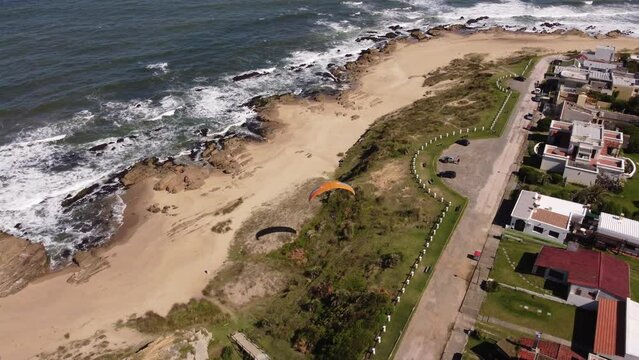 Cinematic drone shot of flying Para-glider over coastline with dunes,sand and ocean waves during summer day - La Pedrera in Uruguay