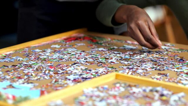 African American woman solving a jigsaw puzzle moves hand over the table and sets down a piece. Hand held closeup clip, with shallow depth of field focus.