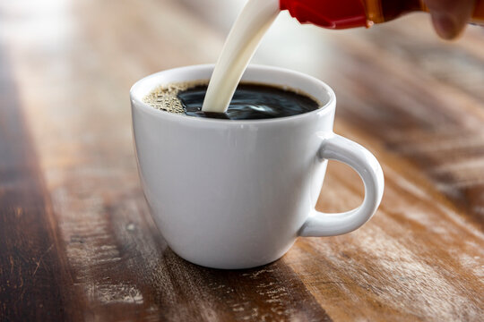 pouring creamer into black coffee in a white mug on a wood table.