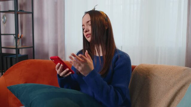 Sad teen girl in sweater use mobile cell phone typing browsing, loses becoming surprised sudden lottery results, bad fortune, loss. Young woman at home in living room on sofa. People sincere emotions