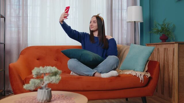Woman resting on sofa at home with smartphone. Pretty smiling female looks at device screen taking selfie on mobile cell phone cam, make virtual video call online on modern application. Social media