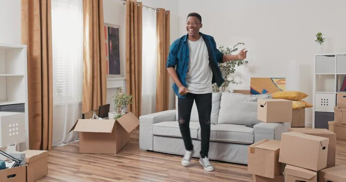 A man is dancing in the middle of the living room in a new apartment guy is happy to move, cardboard boxes with packed things stand around him, joy, smile, a new chapter of life