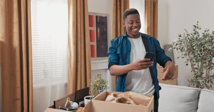 Handsome guy hangs out in new apartment after move-in, unpacks cardboard boxes of packed stuff, writes messages on phone, sends friends selfie brags smiles