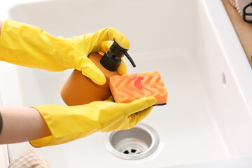 Woman in rubber gloves pouring dish wash detergent onto sponge over sink, closeup