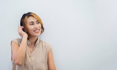 Attractive Asian woman with short hair in beige sleeveless shirt enjoy listening to music, podcast, or song on the radio with white headphone with Happy smile on white background with copy space.