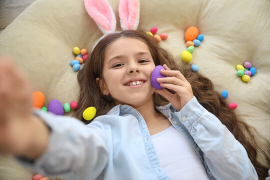 Cute little girl with bunny ears and Easter eggs taking selfie while lying on cushion
