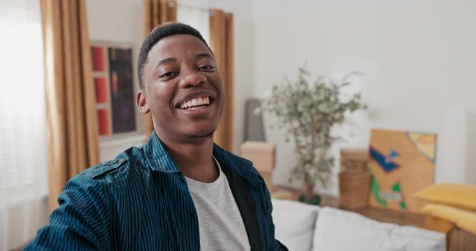 Smiling guy records vlog, video on social media, talks through camera with family man waves hand in greeting shows new apartment cardboard boxes with packed things tells impressions