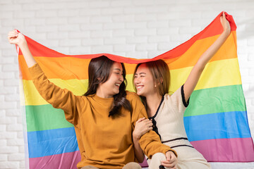 LGBTQ Asian lesbian young couple smiling holding pride flag happiness with love together.Cheerful Homosexual couple embracing and looking at camera under LGBTQ pride flag.LGBTQ Lifestyle Pride Month