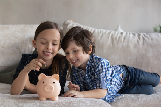 Cheerful sibling little kids putting coin into pink piggybank, playing financial game, resting on couch at home. Children saving money, cash, learning economy, planning future investment.