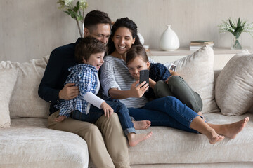 Cheerful family couple and excited happy children resting on sofa, using digital device, taking selfie on smartphone, making video call, looking at mobile phone screen, smiling, laughing, having fun