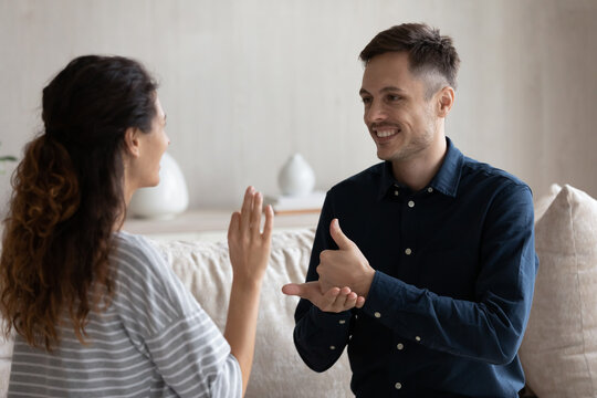Smiling man and woman with deafness using signs for communication, sitting on couch at home, smiling. Therapist teaching gesture language to patient with disability. Hearing disorder concept