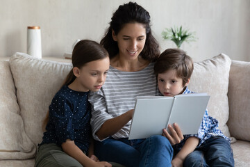 Happy engaged mom reading paper book to son and daughter. Mother and kids sitting on couch at home,...