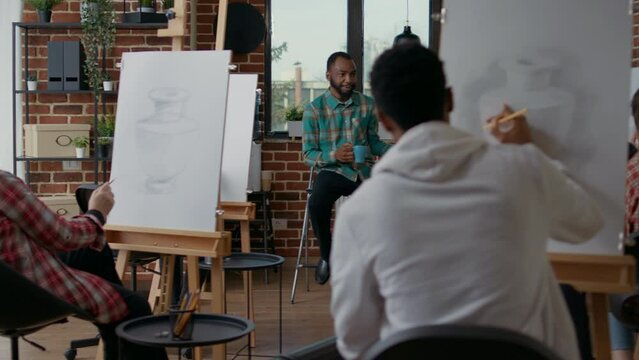 Young man teaching creative people to draw on canvas, explaining drawing technique to practice skills at art class lesson. Teacher talking to students about artistic vision and inspiration.