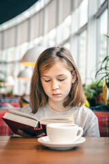 A little girl is reading a book while sitting in a cafe with a cup of tea.