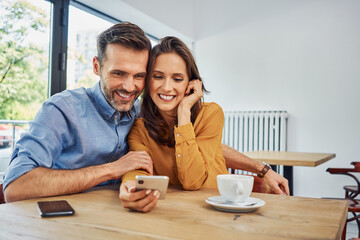 Happy couple looking at smartphone sitting together in cafe browsing together social media