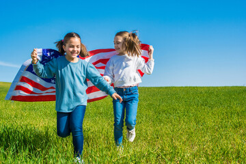 Two Beautiful Girls Running Through A Green Meadow With A Blue Sky, They Are Enjoying The 4th Of July, Showing The Flag Of The United States.