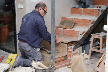 Senior bricklayer building a ceramic wood effect staircase in a home interior. House renewal.