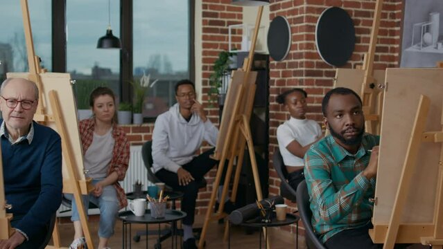 Senior teacher explaining drawing technique to diverse team of students in art class. People using pencil and canvas on easel to develop new artistic skills and to draw vase model on table.