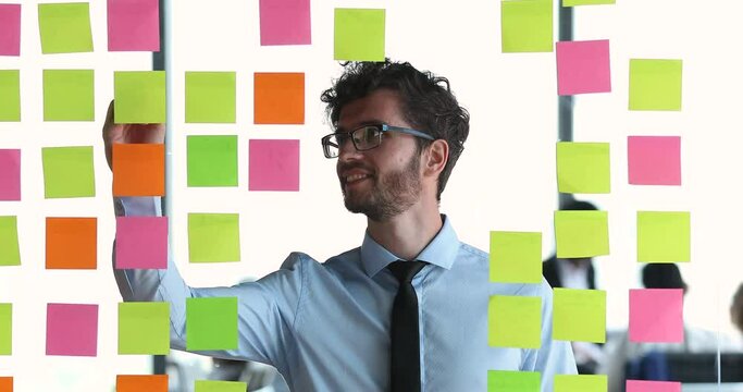 Inspired young businessman entrepreneur executive in glasses mastering kanban scrum agile method to organize and manage data read write notes on sticky cards. View through glass wall in modern office