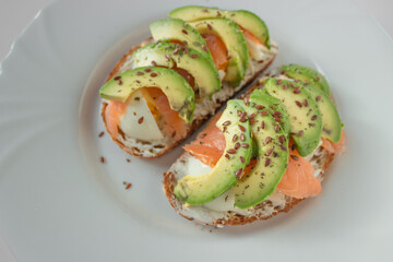 Open sendvich with slices of avocado, fresh salmon, boiled egg and flax seed on a white plate. Healthy eating at home.