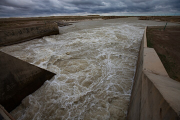 Fototapeta premium Aklak dam, Kzylorda region, Kazakhstan. Concrete channel on Shardara (Syrdarya) river. Turbulent yellow water, grey cloudy sky.