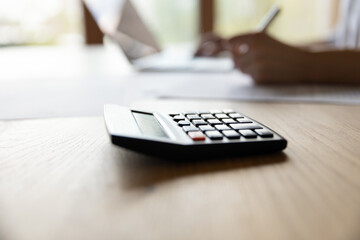 Close up focus on calculator with blurred young woman planning domestic monthly budget alone at home, managing household expenditures, doing financial accounting using e-banking computer application.