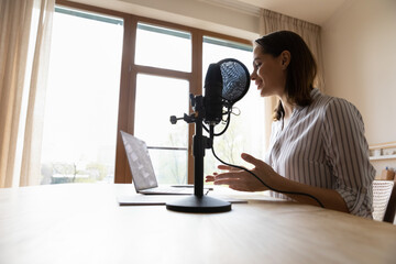 Smiling professional female radio host talking in stand microphone, looking at laptop screen, voice...