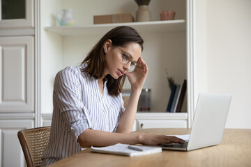 Confused unhappy young Caucasian businesswoman in eyewear looking at laptop screen, feeling stressed stuck with difficult task, considering hard problem solution, overworking in modern home office.