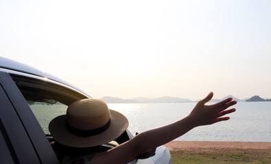 Happy young woman wearing hat waving outside open window car with sea and mountain background. People lifestyle relaxing as traveler on road trip in holiday vacation. Transportation and travel concept