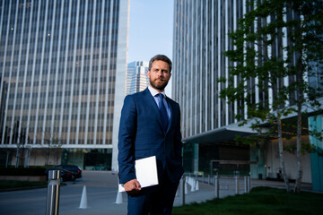 Business man holding laptop standing on the street near business office building.