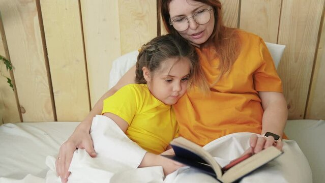 mother in bed reading a book to her daughter. mother and child girl reading a book in bed before going to sleep. happy family kid dream concept indoor lifestyle