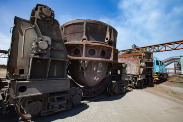 Metal alloys plant (smelter). Blue locomotive and rusted slag car wagons. Pipeline and blue sky on background.