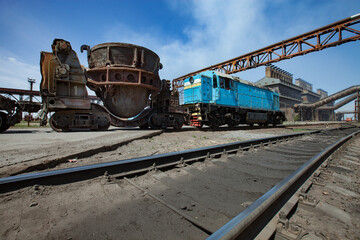 Fototapeta na wymiar Metal alloys plant. Blue locomotive and rusted slag cars. Metallurgical plant (smelter) main industrial building on background. Blue sky.