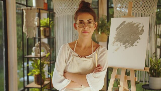 Portrait Of Young Talented Young Female Artist, Wearing Apron, Crosses Arms, Looks At The Camera With A Smile In An Art Studio With Canvas In Sunset, An Freelance Relaxes In Her Free Time After Work