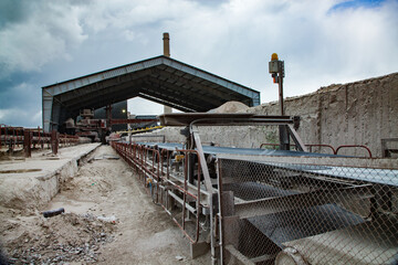 Old Soviet cement plant. Rubber conveyor belt on the rolls. Metal roof and loading machine on background. Blue sky, clouds. Temirtau, Kazakhstan.