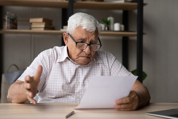 Confused frustrated elder senior 80s man in glasses reading document at table, receiving bad concerning news, getting bank loan notice, feeling puzzled about high insurance fees, doing paperwork