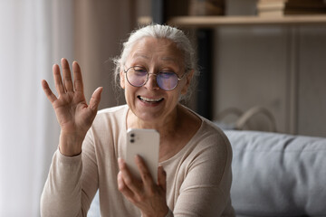 Happy older 70s smartphone user woman waving hand hello at screen, frontal camera, speaking on...