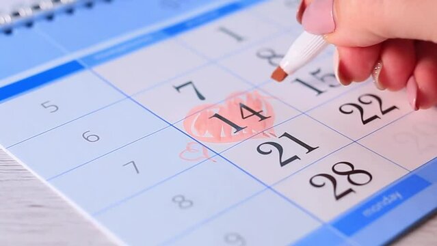 Female hand with red marker paint a heart shape in the calendar february 14th near gift. Valentine's day
