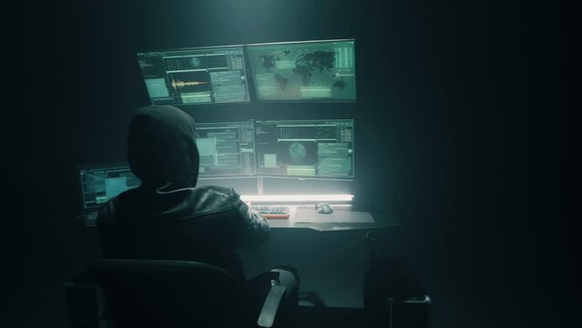 Zoom in and zoom out pan around view of man in hoodie typing on computer keyboard, while preforming cyber attack under spotlight in dark room on companys servers