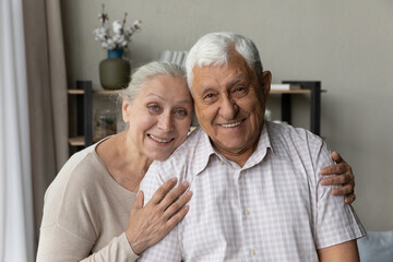 Happy elderly 70s 80s couple posing in home interior head shot portrait. Old grey haired husband and wife hugging with love, tenderness, care, looking at camera together with toothy smiles, laughing