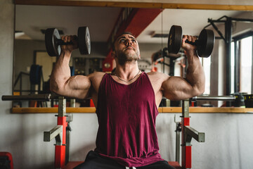 Front view of one adult man male bodybuilder doing shoulder press exercise with dumbbells gym...