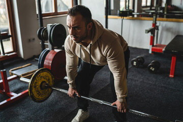 Fototapeta na wymiar One man young adult caucasian male bodybuilder training back doing deadlift with barbel and weights while standing in the gym wearing hoodie real people copy space side view full length