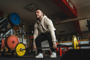 Fototapeta na wymiar One man young adult caucasian male bodybuilder training back doing deadlift with barbel and weights while standing in the gym wearing hoodie real people copy space side view full length