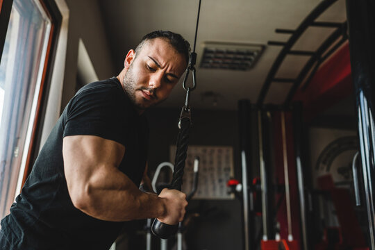 One man side view caucasian male bodybuilder at gym wearing black shirt using rope and cable weights for arms exercise muscular triceps training copy space waist up dark photo real people bodybuilding