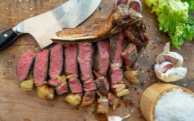 Sliced sous vide cooked medium-rare dry aged rubia gallega beef steak with knife, sea salt flakes, garlic, pepper and salad on a wooden board