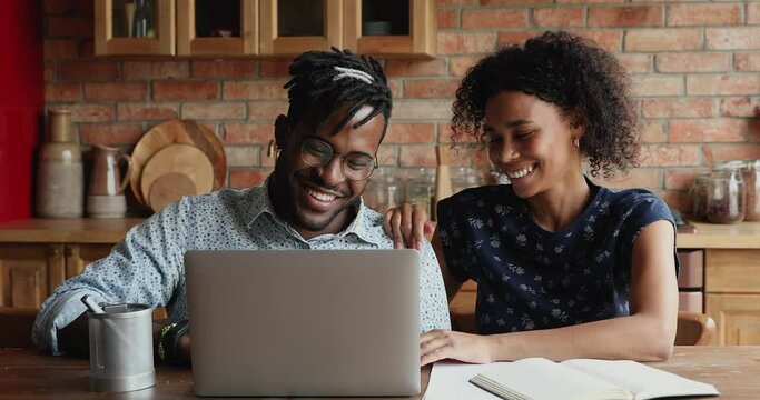 Happy smiling young African married couple sit by laptop surf social media chat discuss funny online news. Loving young wife hug shoulder of dear husband using pc having fun talking at home kitchen - Powered by Adobe